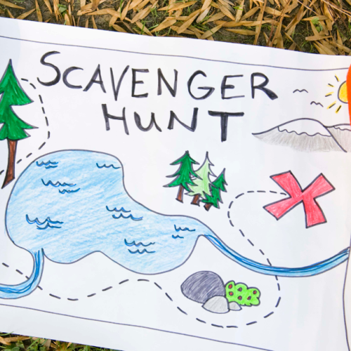 A hand-drawn scavenger hunt map on grass features a lake, trees, a mountain, and a red X, suggesting a treasure location.