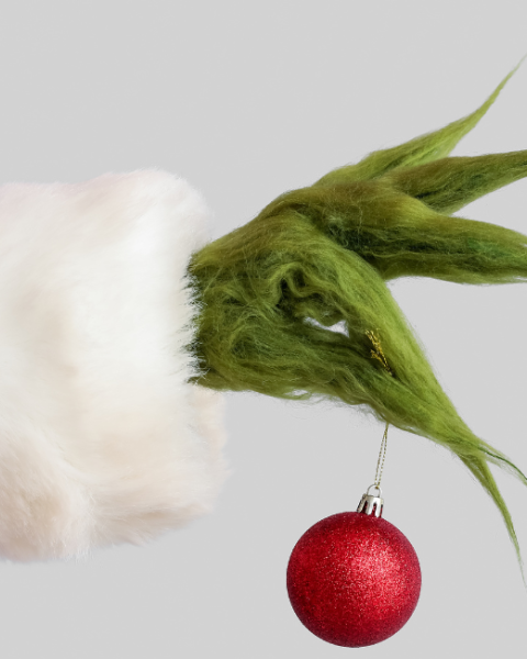 A hairy green hand in a red Santa sleeve holds a red Christmas ornament, suggesting a mischievous holiday character.