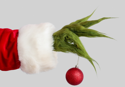 A hairy green hand in a red Santa sleeve holds a red Christmas ornament, suggesting a mischievous holiday character.