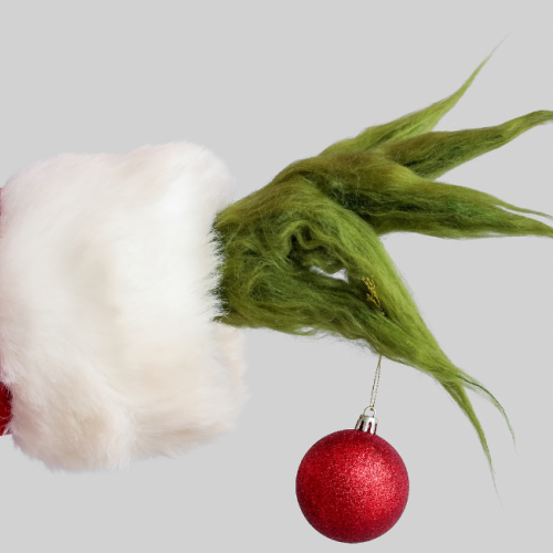 A hairy green hand in a red Santa sleeve holds a red Christmas ornament, suggesting a mischievous holiday character.