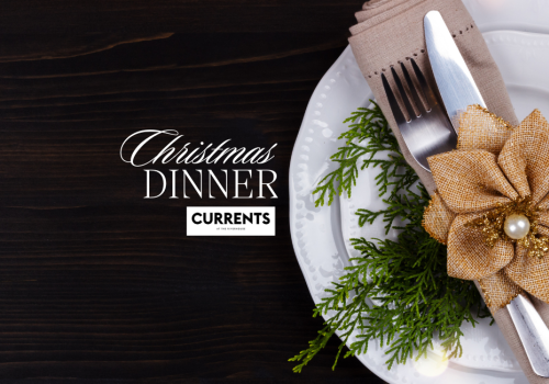 Place setting with a knife and fork wrapped in a napkin, decorated with a gold flower and greenery, on a plate. Text: Christmas Dinner Currents.