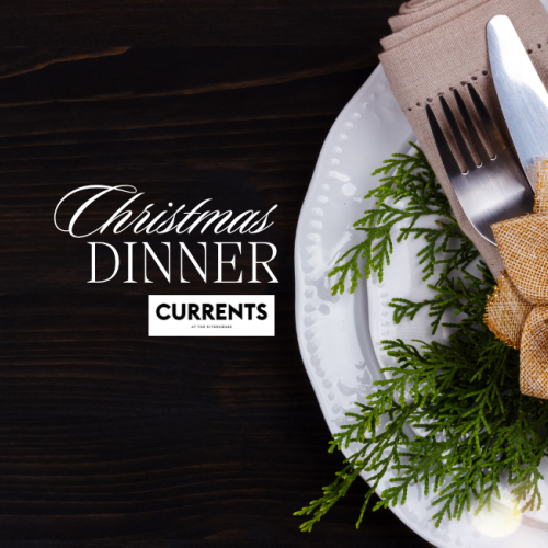 Place setting with a knife and fork wrapped in a napkin, decorated with a gold flower and greenery, on a plate. Text: Christmas Dinner Currents.