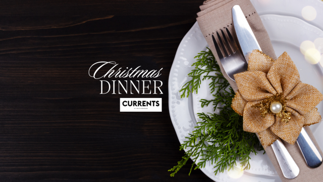Place setting with a knife and fork wrapped in a napkin, decorated with a gold flower and greenery, on a plate. Text: Christmas Dinner Currents.