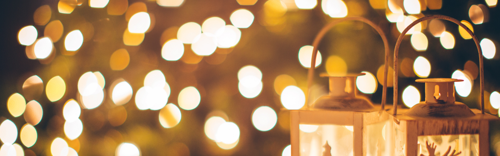 Two decorative lanterns with festive cutouts of a tree and a reindeer, set against a warm, blurry background of holiday lights.