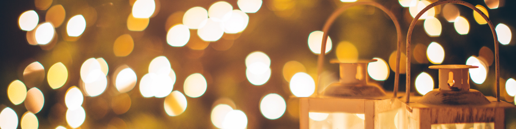 Two decorative lanterns with festive cutouts of a tree and a reindeer, set against a warm, blurry background of holiday lights.