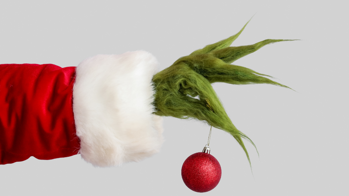 A green, furry hand in a Santa suit holds a red round ornament against a gray background.