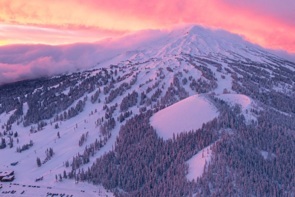 A snow-covered mountain with ski trails under a vibrant pink and orange sky at sunset, surrounded by snow-dusted trees and buildings.