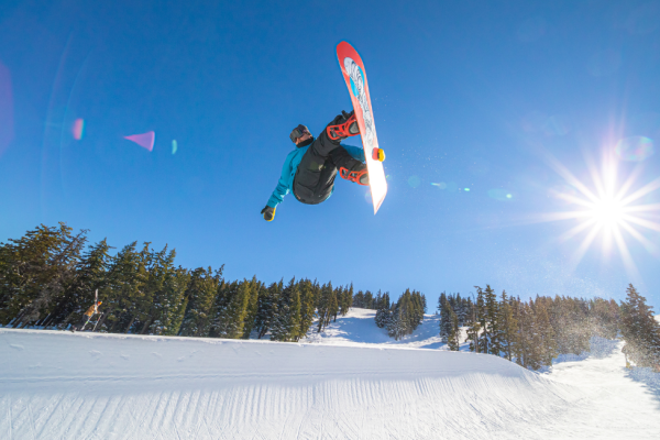 A snowboarder is performing a jump in mid-air on a sunny day, with a backdrop of snow and evergreen trees.