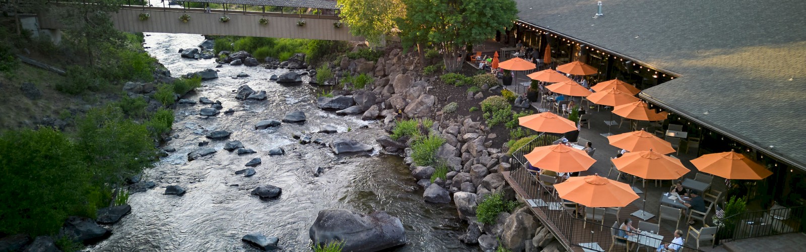 A riverside scene with a building, outdoor seating with orange umbrellas, surrounded by trees and rocks.