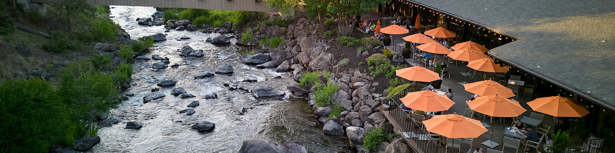 A riverside scene with a building, outdoor seating with orange umbrellas, surrounded by trees and rocks.