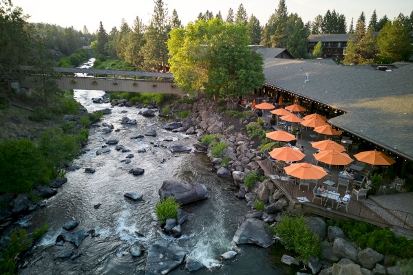 A scenic riverside view shows a building with orange umbrellas on a patio, surrounded by trees and rocks, with a bridge in the background.