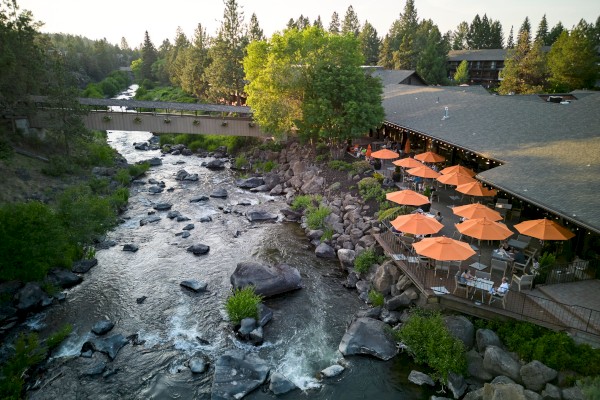 A riverside view with a building featuring outdoor seating and orange umbrellas, surrounded by trees and a wooden bridge.