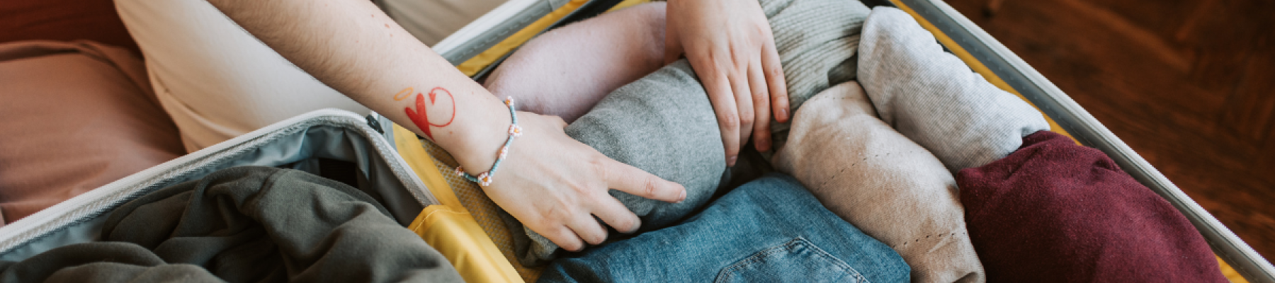 A person is packing a suitcase with neatly folded and rolled clothes, including sweaters and jeans, while wearing bracelets.