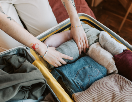 A person is packing a suitcase with neatly folded and rolled clothes, including sweaters and jeans, while wearing bracelets.