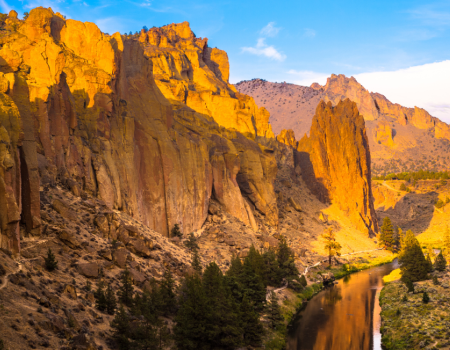 A dramatic landscape with towering orange cliffs illuminated by sunlight, a winding river below, and scattered greenery throughout.