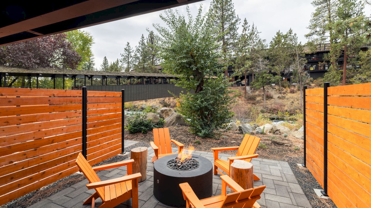 A cozy outdoor patio with four orange chairs around a round fire pit, surrounded by wooden fences and trees in the background.