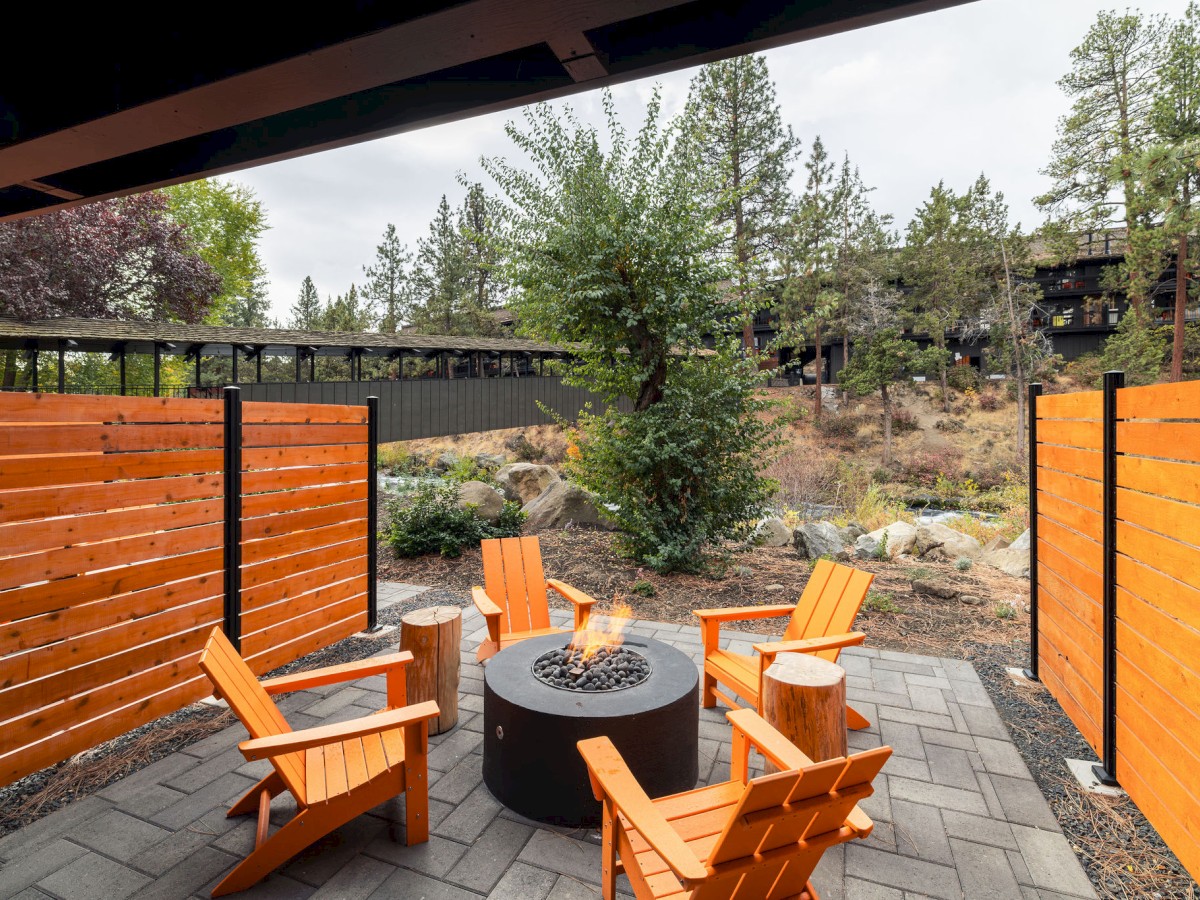 A cozy outdoor patio with four orange chairs around a round fire pit, surrounded by wooden fences and trees in the background.