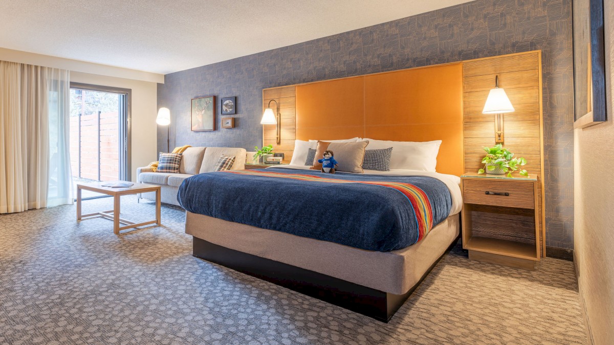 A modern hotel room with a large bed, stylish headboard, sofa, and nightstands. Natural light enters through a window.