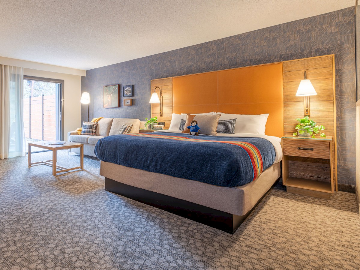 A modern hotel room with a large bed, stylish headboard, sofa, and nightstands. Natural light enters through a window.