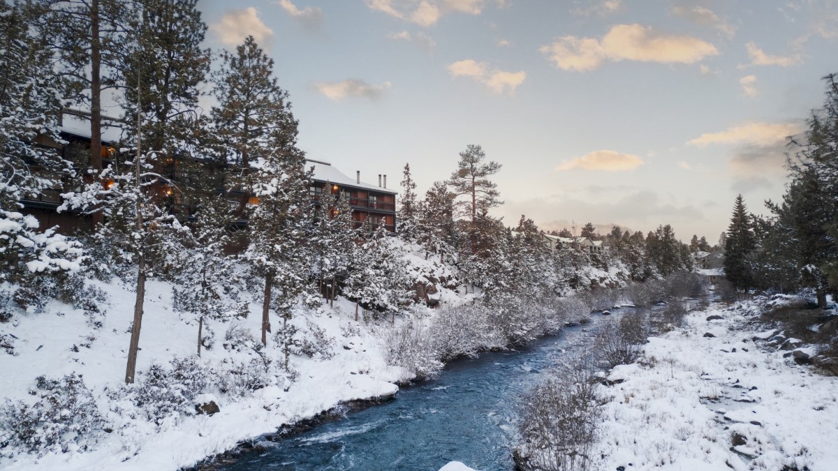 Snow-covered landscape with a river running through a forest. Pines border a cabin under a partly cloudy sky.