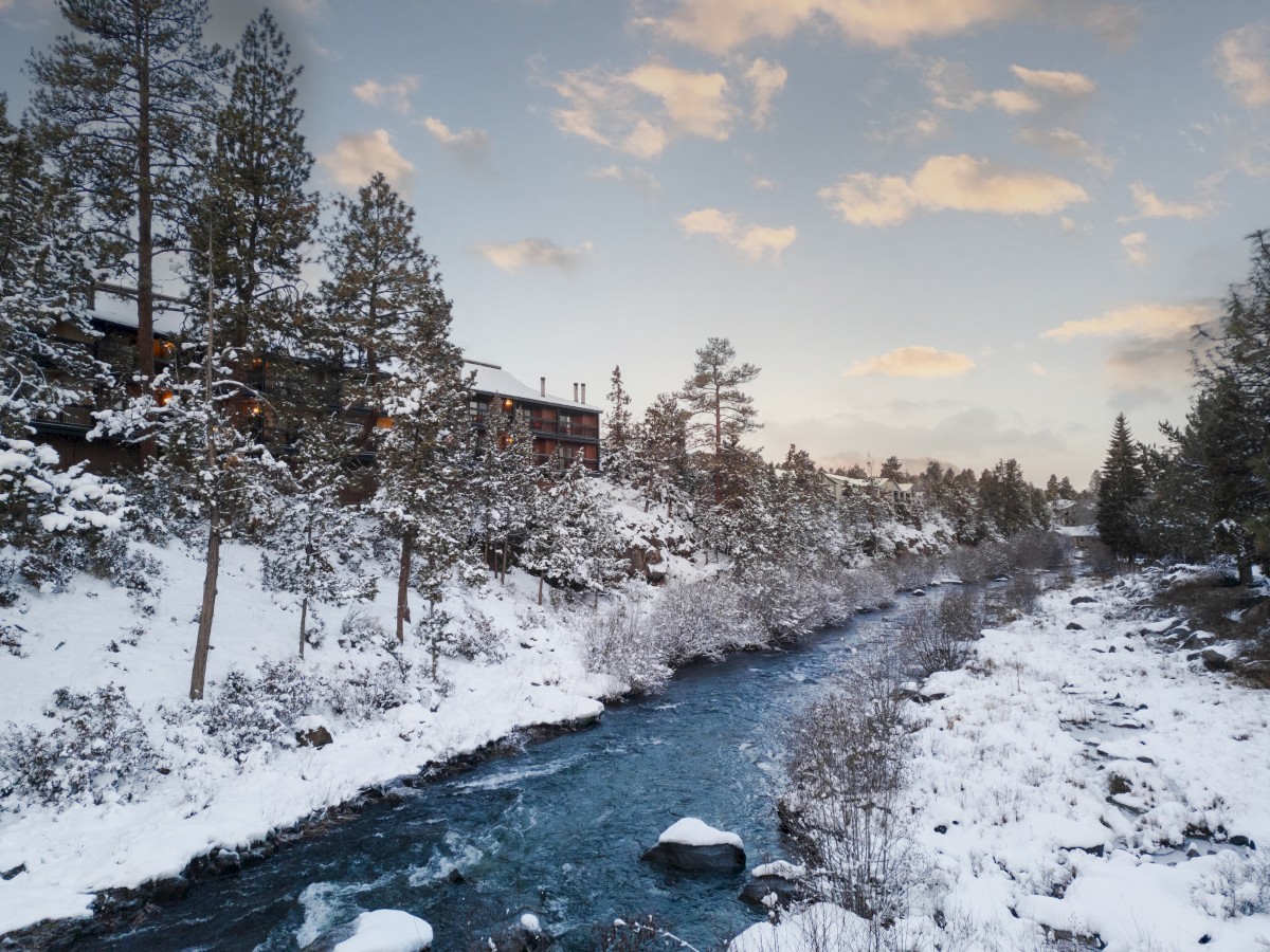 Snow-covered landscape with a river running through a forest. Pines border a cabin under a partly cloudy sky.