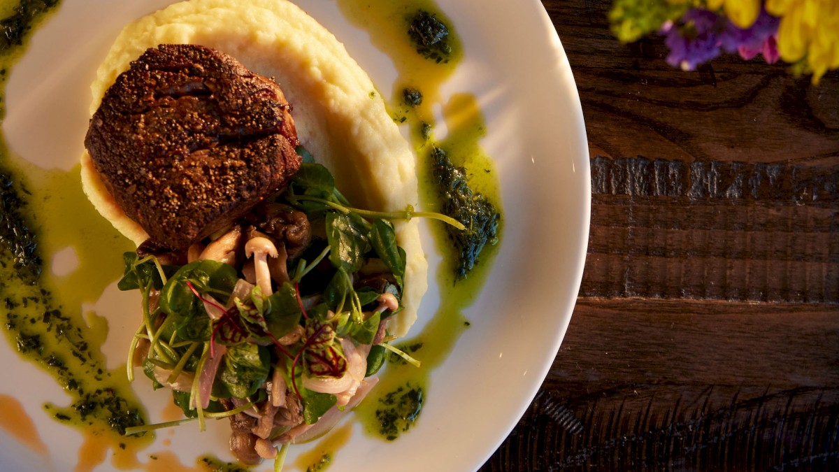 A plate with a steak, mashed potatoes, and greens, garnished with sauce. A glass of white wine and yellow flowers beside it.