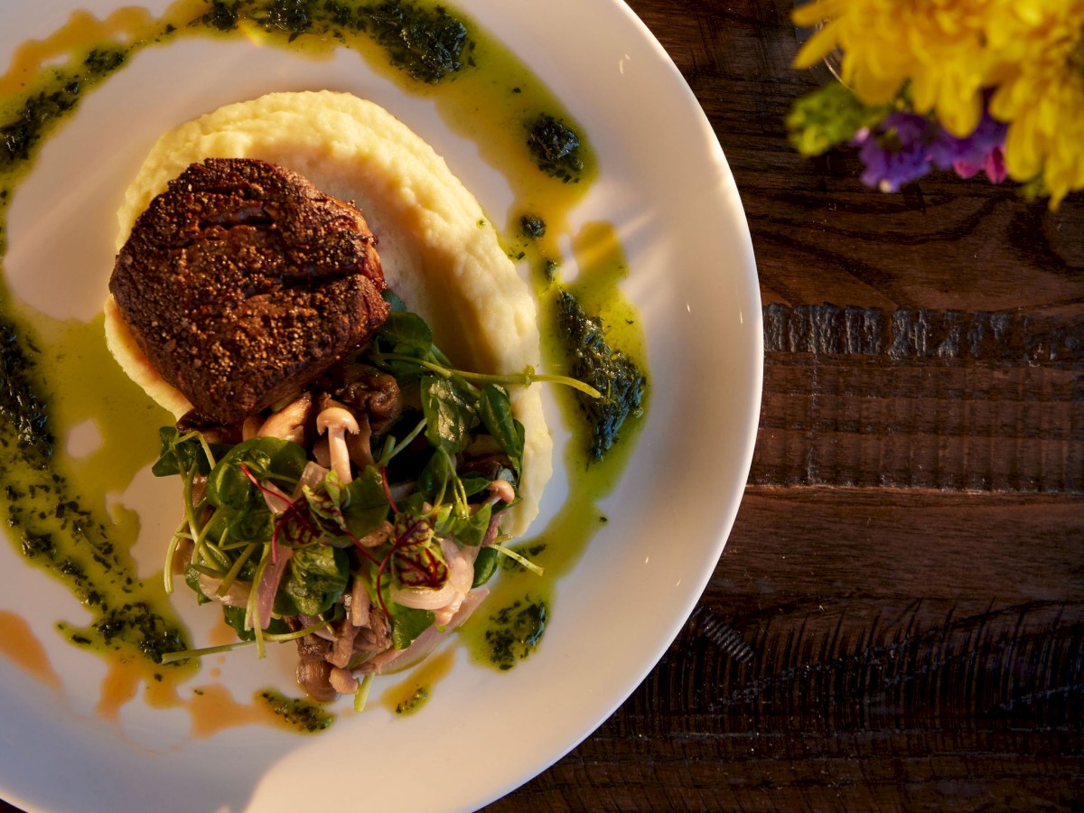 A plate with a steak, mashed potatoes, and greens, garnished with sauce. A glass of white wine and yellow flowers beside it.