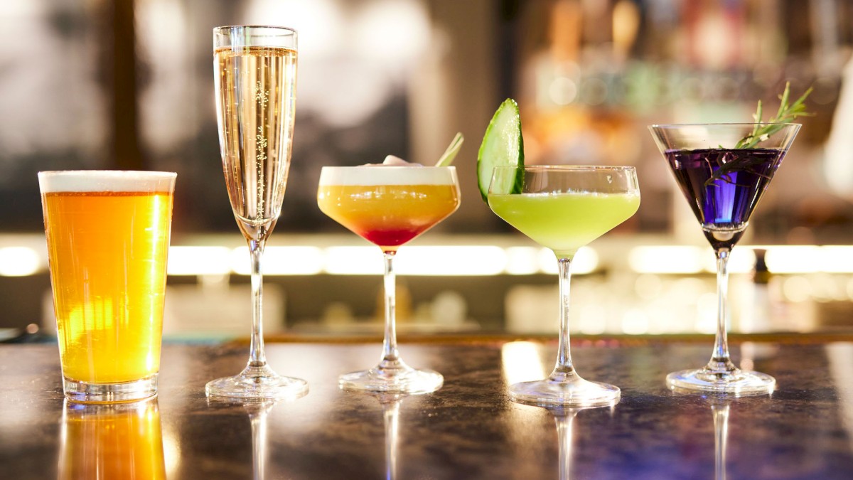 The image shows five different drinks on a bar: a beer, champagne, and three colorful cocktails in various glasses.