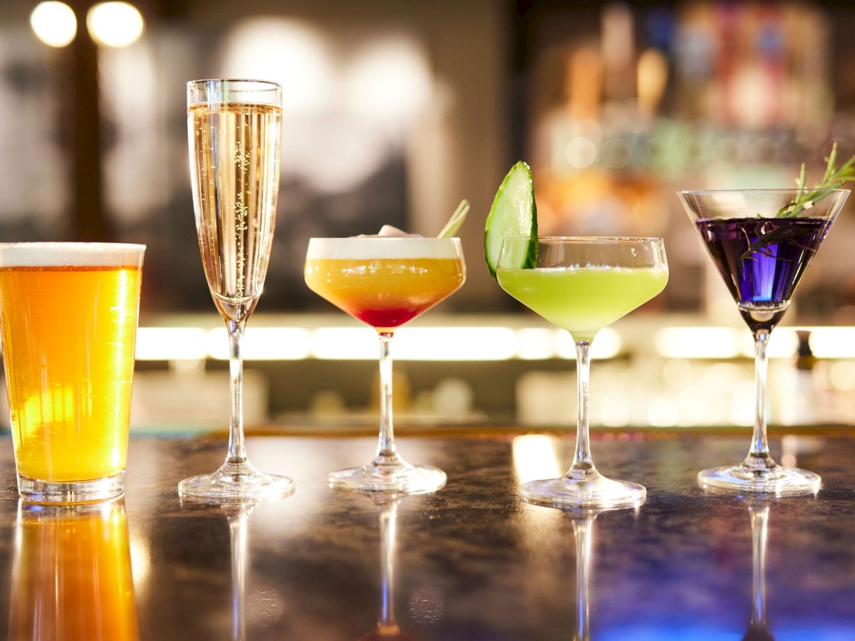 The image shows five different drinks on a bar: a beer, champagne, and three colorful cocktails in various glasses.