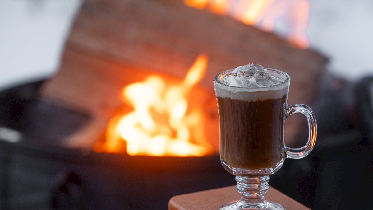 A mug of hot beverage with whipped cream, sitting near a fire pit with burning wood in the background.