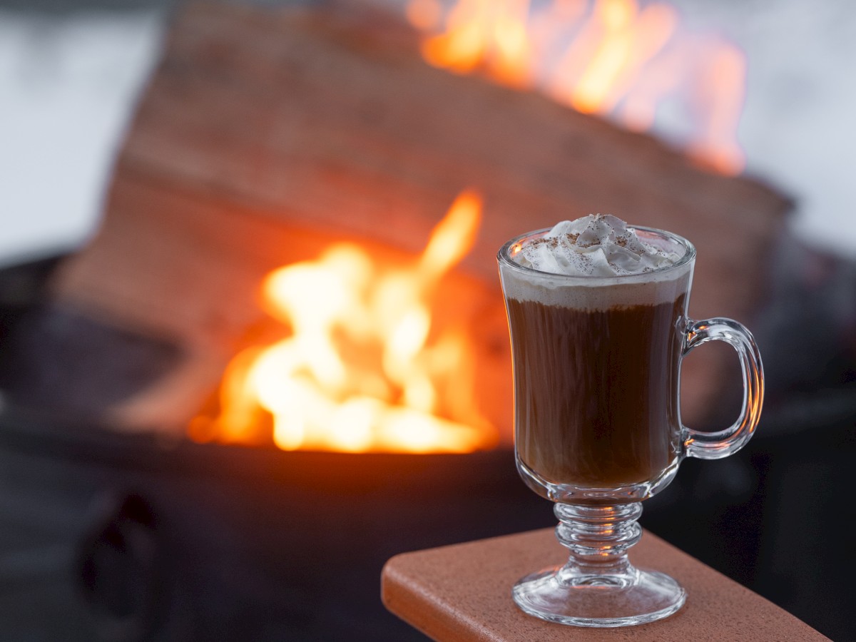 A mug of hot beverage with whipped cream, sitting near a fire pit with burning wood in the background.