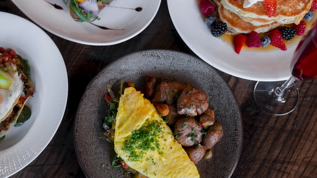 The image shows a variety of breakfast dishes, including an omelette with potatoes, pancakes with berries, and a beverage.