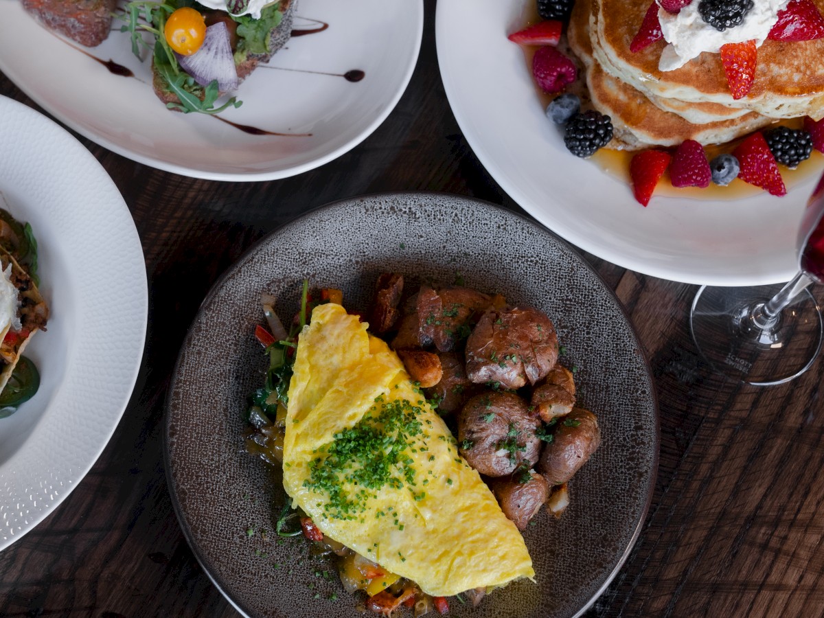 The image shows a variety of breakfast dishes, including an omelette with potatoes, pancakes with berries, and a beverage.