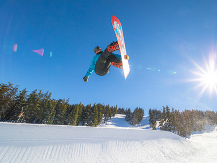 A snowboarder performs a high jump against a clear blue sky, with the sun shining brightly and snow-covered trees in the background.