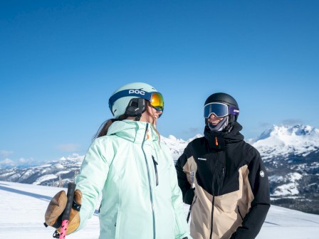 Two snowboarders stand on a snowy mountain ridge in bright jackets, helmets, and goggles, with a blue sky and alpine peaks in the background.