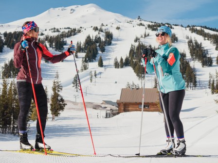 Two people are enjoying cross-country skiing on a snowy landscape with a mountain backdrop. They're dressed in winter gear and holding ski poles.