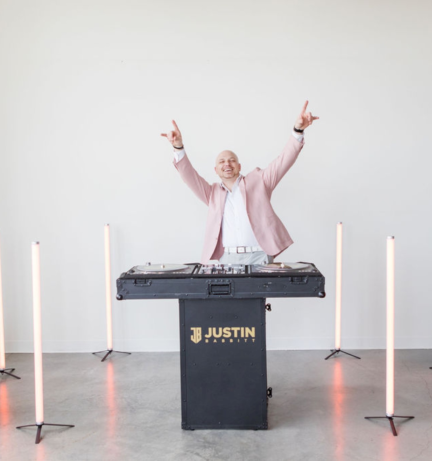 A person in a pink blazer stands behind a DJ booth labeled 