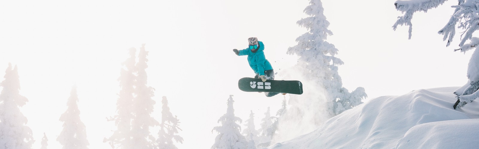 A snowboarder in a blue jacket leaps off a snowy hill, surrounded by snow-covered trees, under a bright sky.