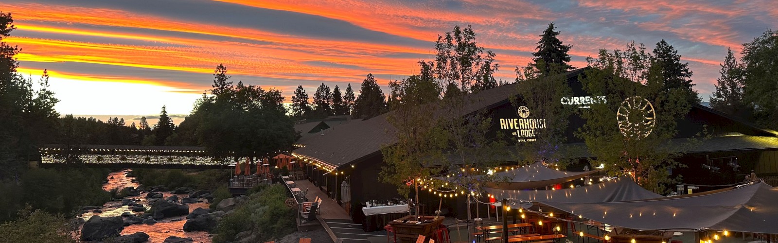 A beautifully lit outdoor dining area beside a river during a vibrant sunset, with trees and a colorful sky in the background.