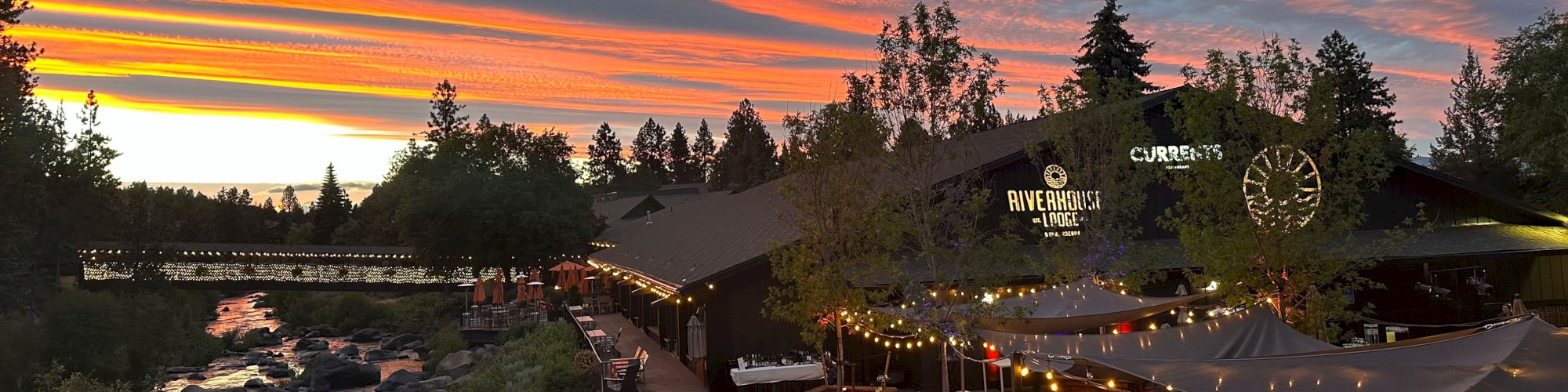 A beautifully lit outdoor dining area beside a river during a vibrant sunset, with trees and a colorful sky in the background.