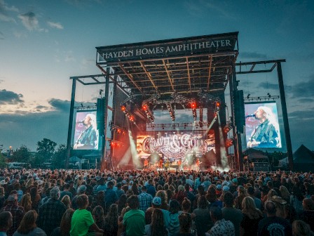 A live concert is taking place at the Hayden Homes Amphitheater, with a large crowd gathered and vibrant stage lighting.
