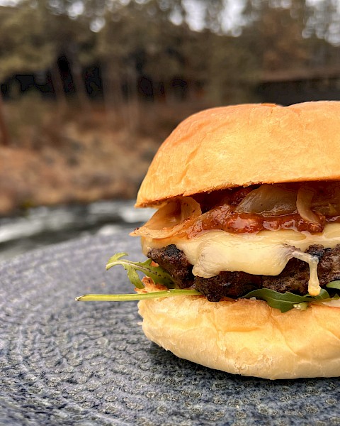 A cheeseburger with bacon, lettuce, and onions on a bun, placed on a textured plate outdoors near a river and trees.