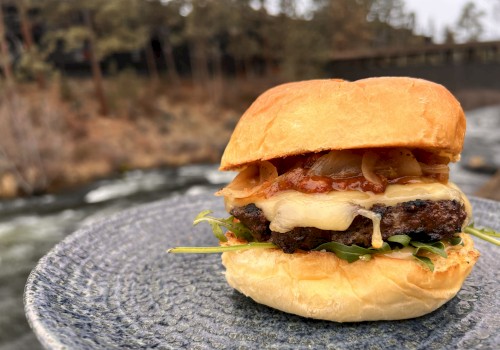 A cheeseburger with bacon, lettuce, and onions on a bun, placed on a textured plate outdoors near a river and trees.
