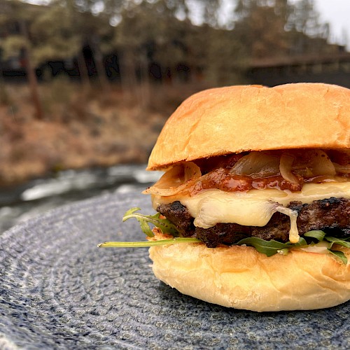 A cheeseburger with bacon, lettuce, and onions on a bun, placed on a textured plate outdoors near a river and trees.