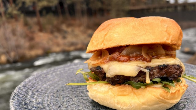 A cheeseburger with bacon, lettuce, and onions on a bun, placed on a textured plate outdoors near a river and trees.