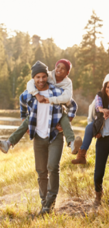 A family is enjoying a walk in a scenic outdoor setting, with adults giving piggyback rides to children, under a clear, sunny sky.