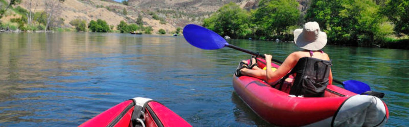 Two people kayaking on a calm river surrounded by hills and greenery, under a clear blue sky.