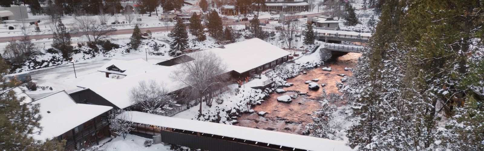 Aerial winter view of Riverhouse Lodge along the Deschutes River in Bend, Oregon, showcasing snow-covered grounds, a covered footbridge, and scenic river views while promoting the Hot Dates = Hot Rates limited-time hotel offer for short-term travel.