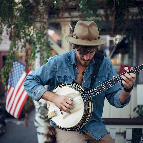 Musician, Oyster Lee, plays banjo on a porch during Father’s Day at Currents at Riverhouse Lodge in Bend, Oregon, with greenery and an American flag in the background.