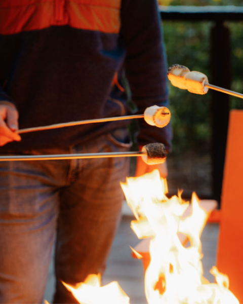 Guest roasting marshmallows over a fire pit at Riverhouse Lodge with signature orange chairs and Deschutes River views in the background during Stars & S’mores event.